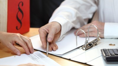 Beratung: ein Mann erläutert einem anderen Mann ein Formular (nah, nur Hände, Formular, aufgeschlagener Aktenordner, Taschenrechner und ein Buch mit einem aufgedruckten Paragraphenzeichen sind sichtbar)
